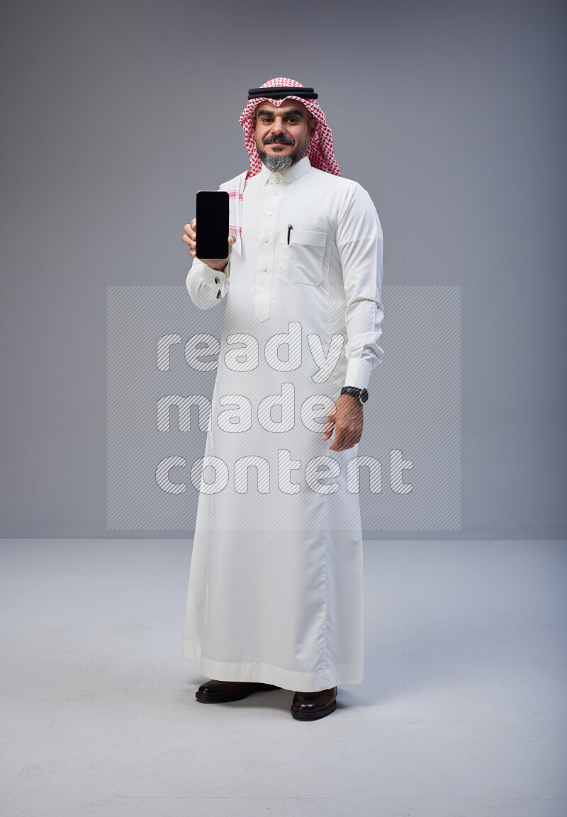 Saudi man Wearing Thob and red Shomag standing showing phone to camera on Gray background