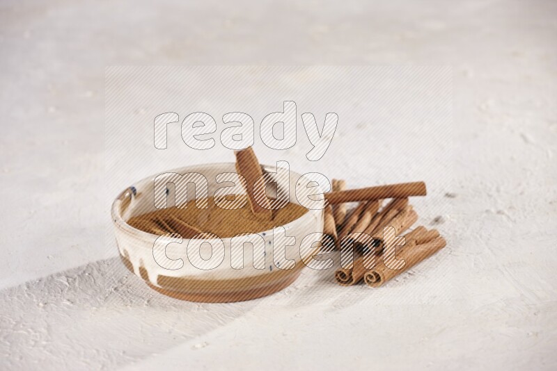 Ceramic bowl full of cinnamon powder with cinnamon sticks on the side on white background