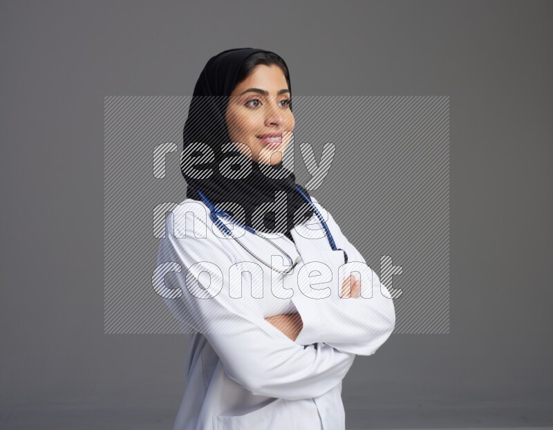 Saudi woman wearing lab coat with stethoscope standing with crossed arms on Gray background