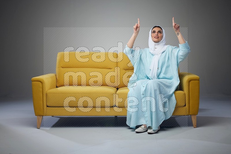 A Saudi woman wearing a light blue Abaya and white head scarf sitting on a yellow sofa and thinking and pointing up eye level on a grey background