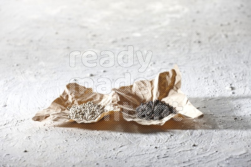 2 Crumpled pieces of paper full of black and white pepper beads on a textured white flooring