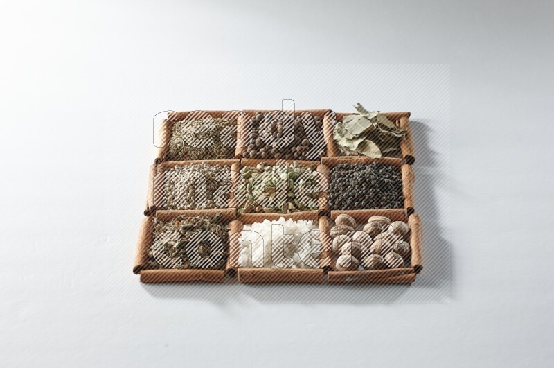 9 مربعات من أعواد القرفة ممتلئة بالهيل في المنتصف محاطة بجوزة الطيب والملح والريحان المجفف والفلفل الأبيض والكمون وبهار الفلفل الحلو وأوراق الغار والفلفل الأسود على أرضية بيضاء