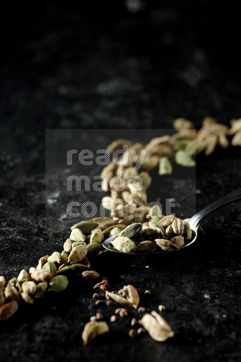 A Metal spoon full of cardamom seeds and some seeds beside it on a textured black flooring