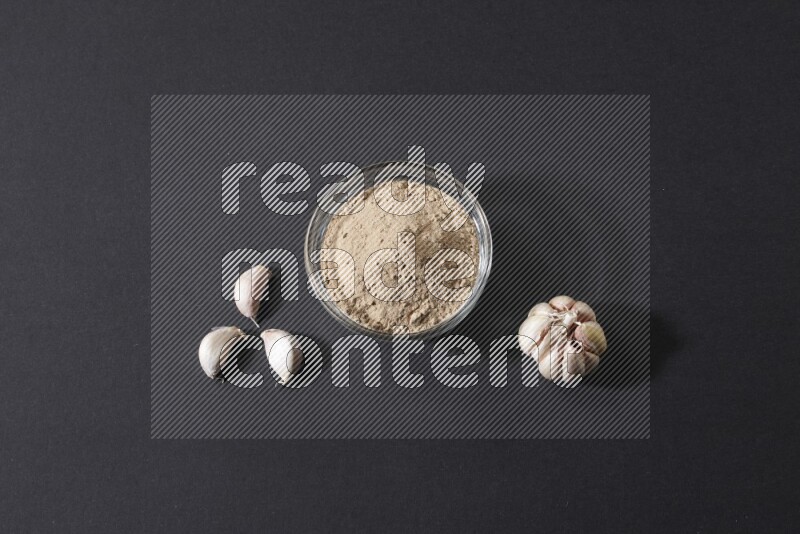 A glass bowl full of garlic powder with garlic bulb and some cloves beside it on a black flooring