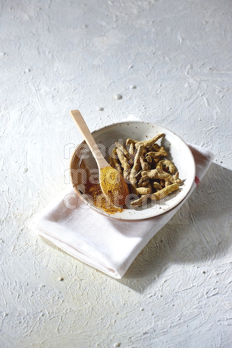A plate filled with dried turmeric whole fingers and a wooden spoon full of turmeric powder on a textured white flooring