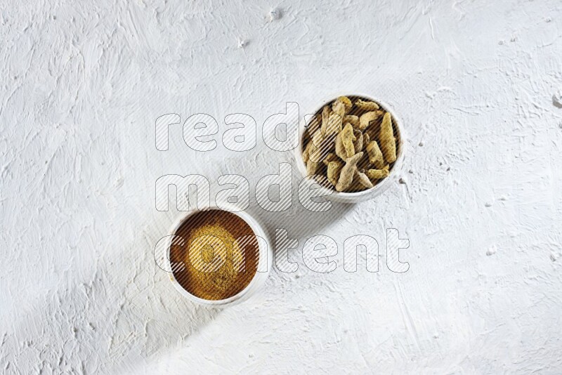 وعاءان فخاريان بيج ممتلئان ببودرة الكركم وأصابع الكركم المجففة على خلفية بيضاء