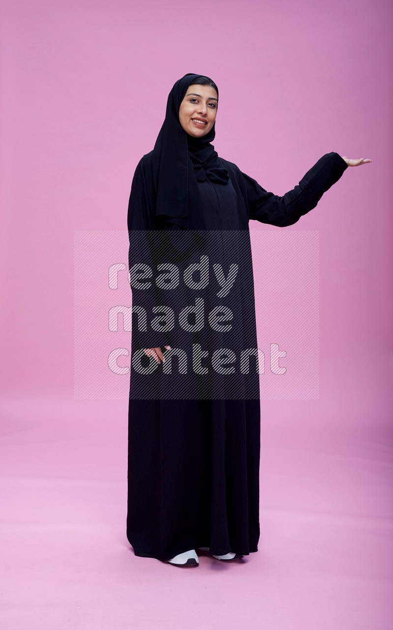 Saudi woman wearing Abaya standing interacting with the camera on pink background