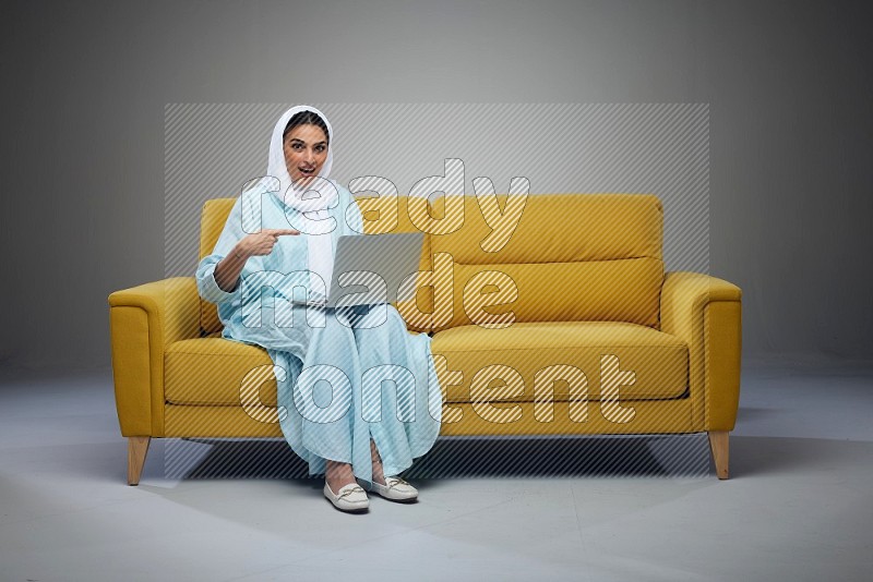 A Saudi woman sitting on a yellow Sofa With her Laptop Pointing wearing Blue Abaya
