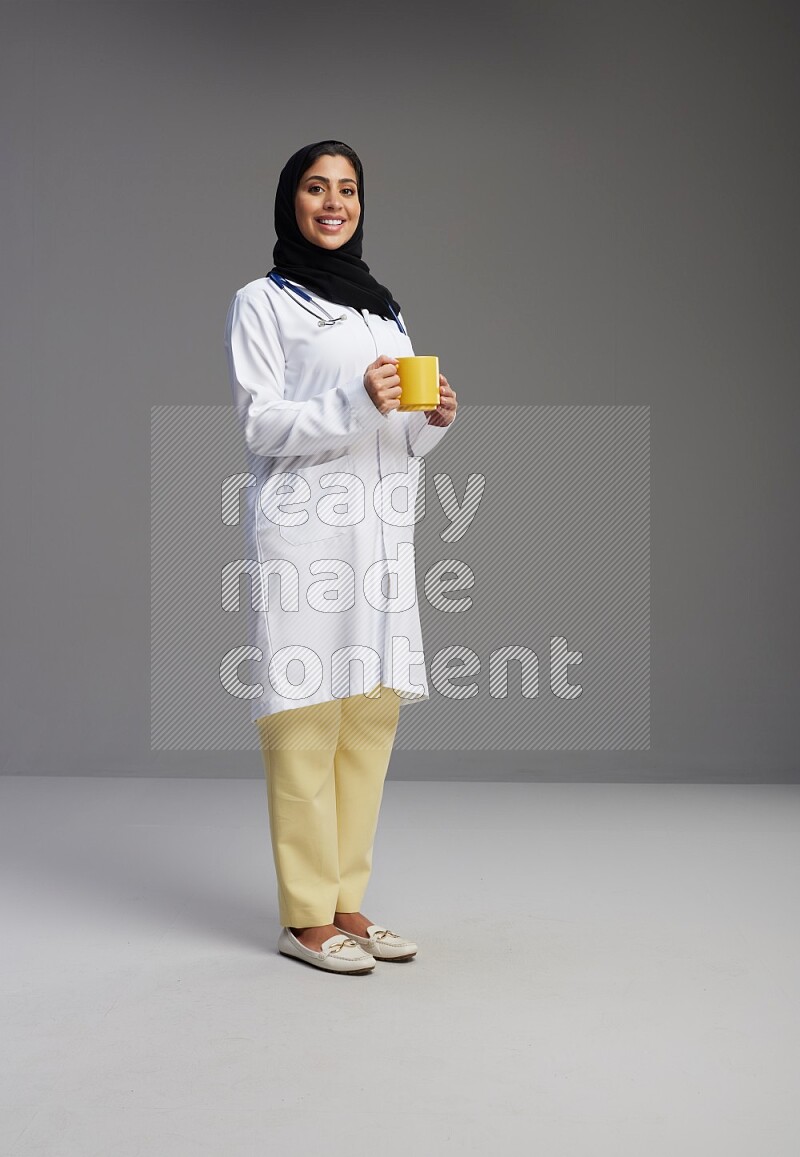 Saudi woman wearing lab coat with stethoscope standing holding mug on Gray background
