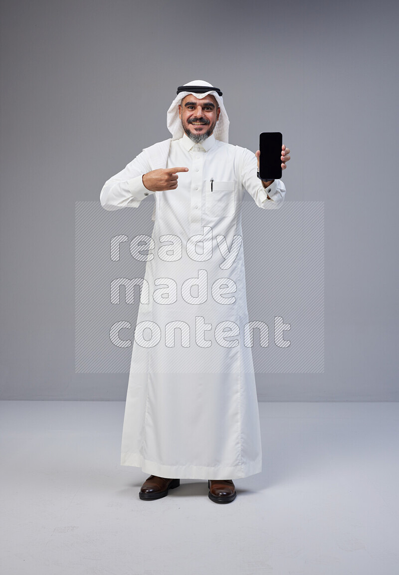 Saudi man Wearing Thob and white Shomag standing showing phone to camera on Gray background