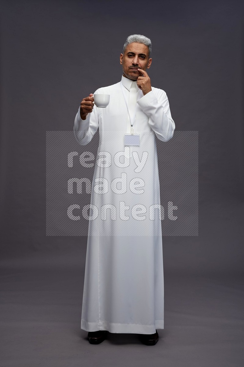Saudi man wearing thob with neck strap employee badge standing holding mug on gray background