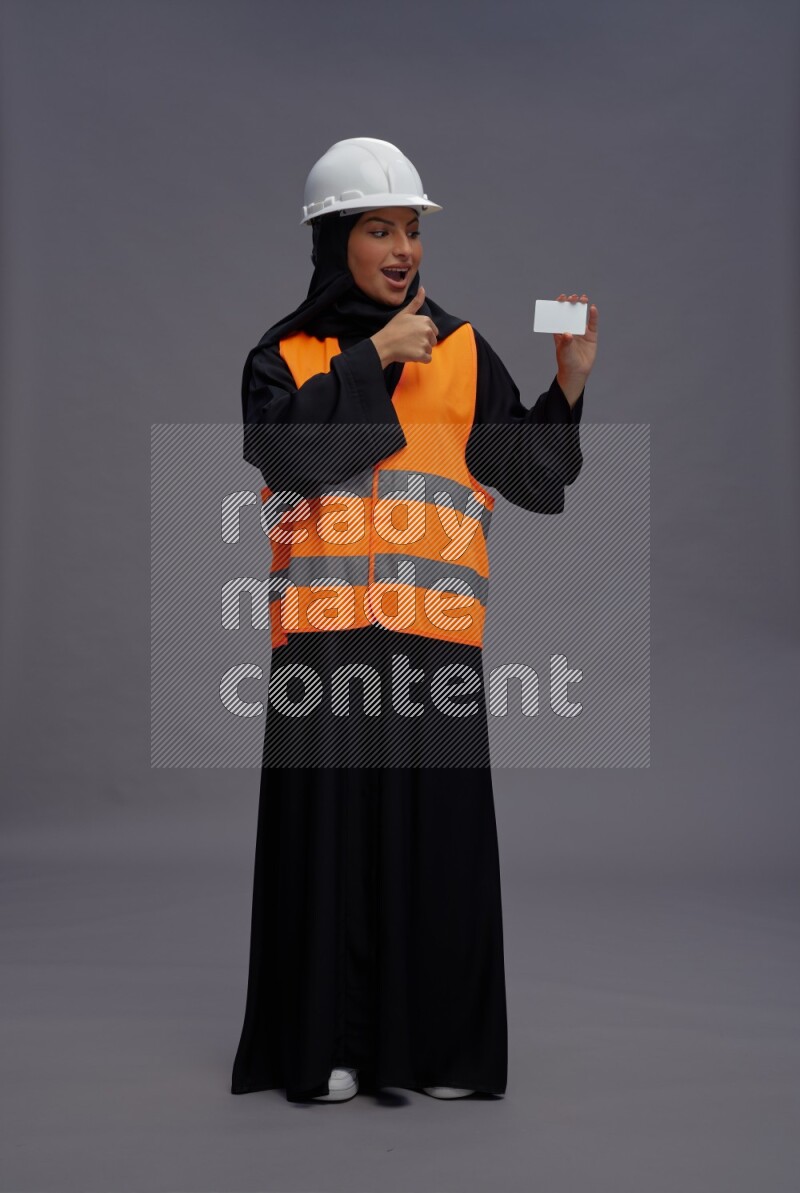 Saudi woman wearing Abaya with engineer vest standing holding ATM card on gray background