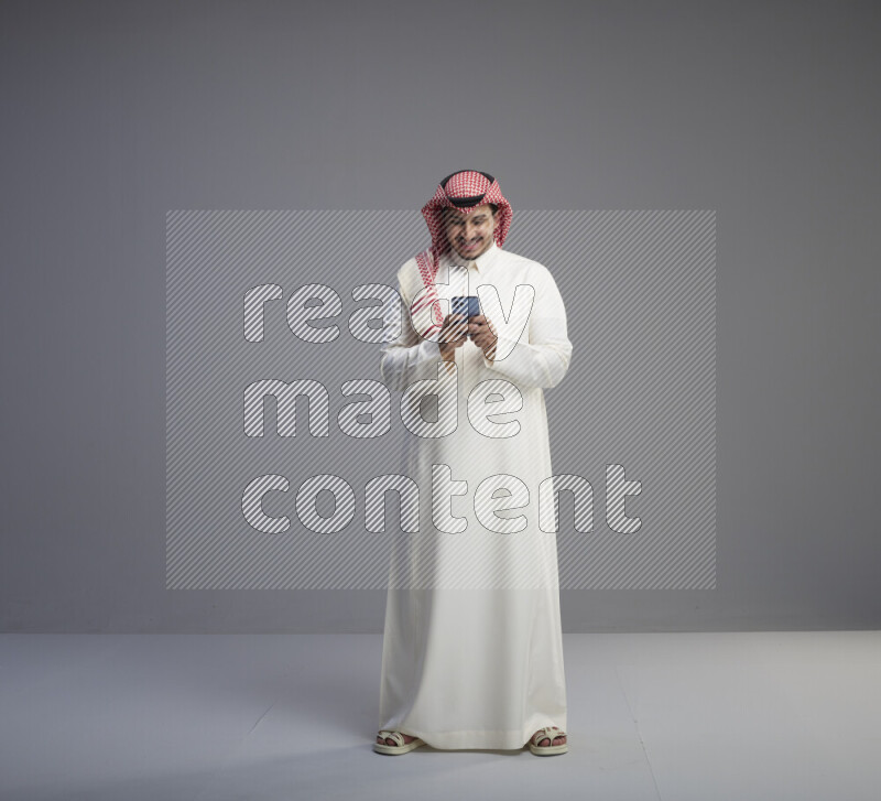A  Saudi man standing wearing thob and red shomag texting on phone on gray background