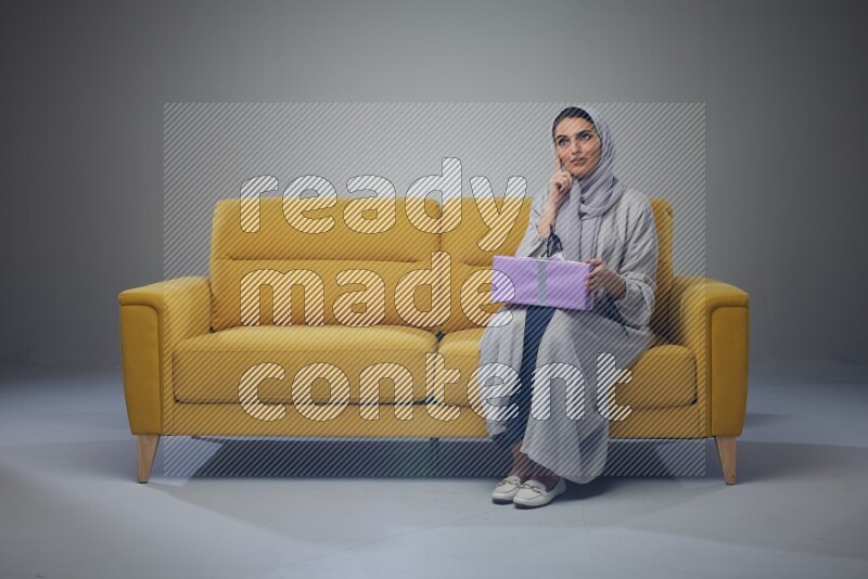 A Saudi woman wearing a light gray Abaya and head scarf sitting on a yellow sofa holding a gift box beside multi color gift boxes eye level on a grey background