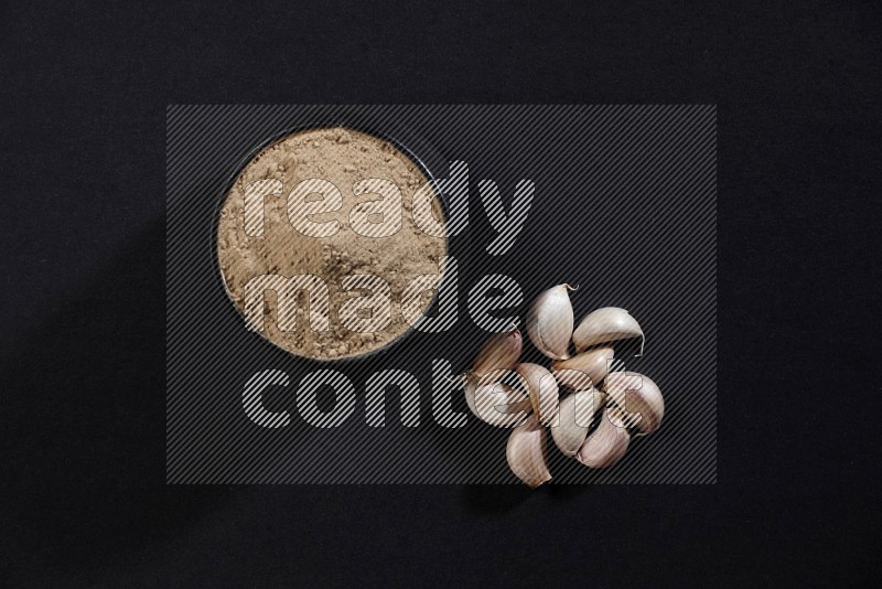 A black pottery bowl full of garlic powder and cloves beside it on a black flooring in different angles