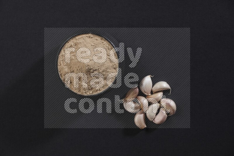 A black pottery bowl full of garlic powder with some cloves beside it on a black flooring