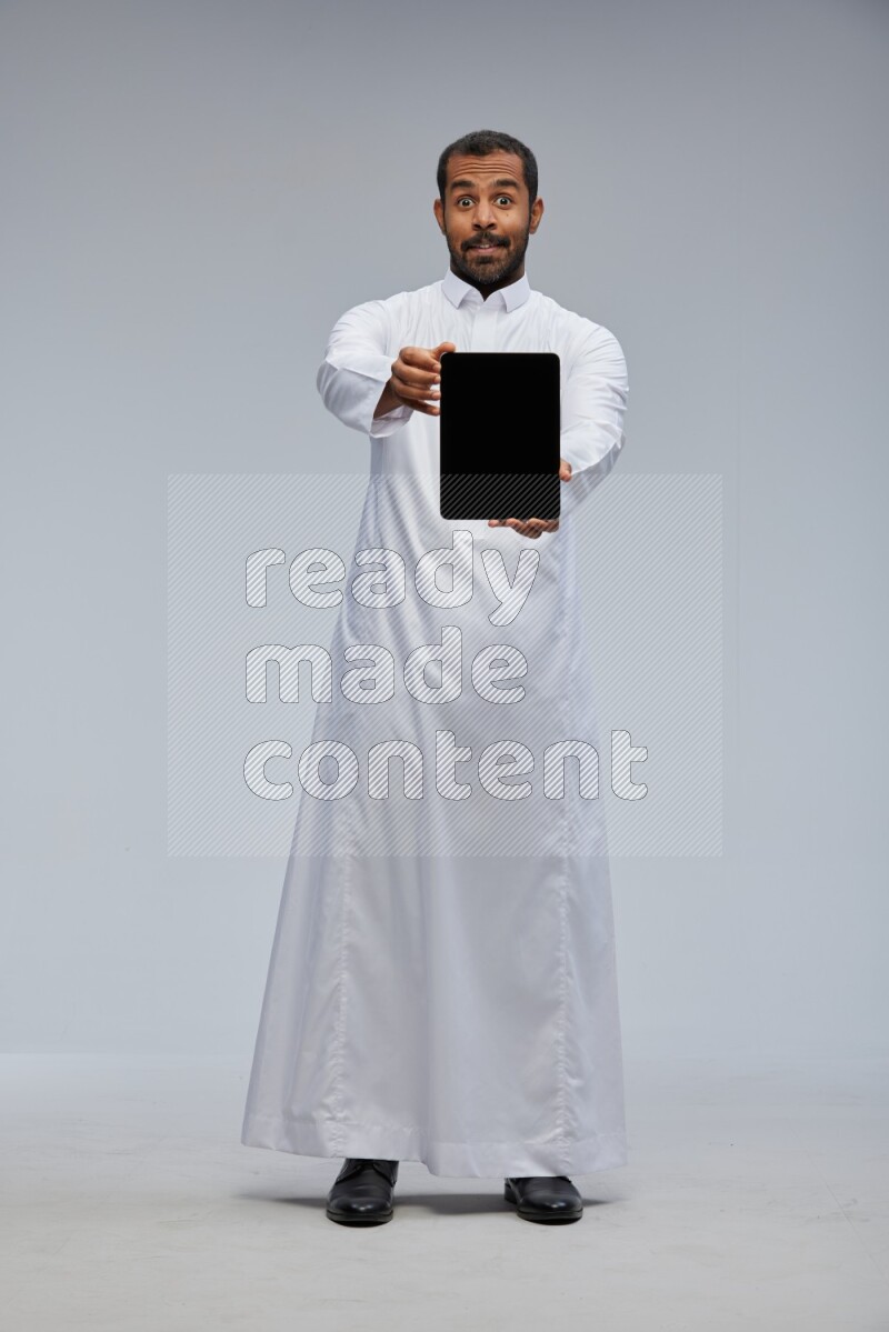 Saudi man wearing Thob standing showing tablet to camera on Gray background