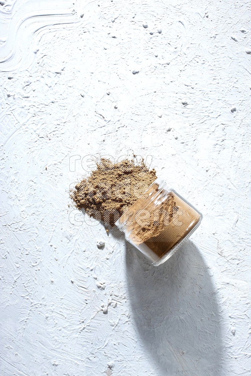 A flipped glass jar full of allspice powder and powder spilled out of it on a textured white flooring