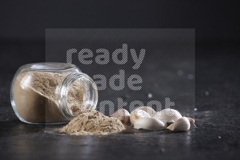 A glass spice jar full of garlic powder flipped over with the powder came out on a textured black flooring