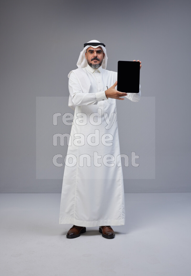 Saudi man Wearing Thob and white Shomag standing showing tablet to camera on Gray background