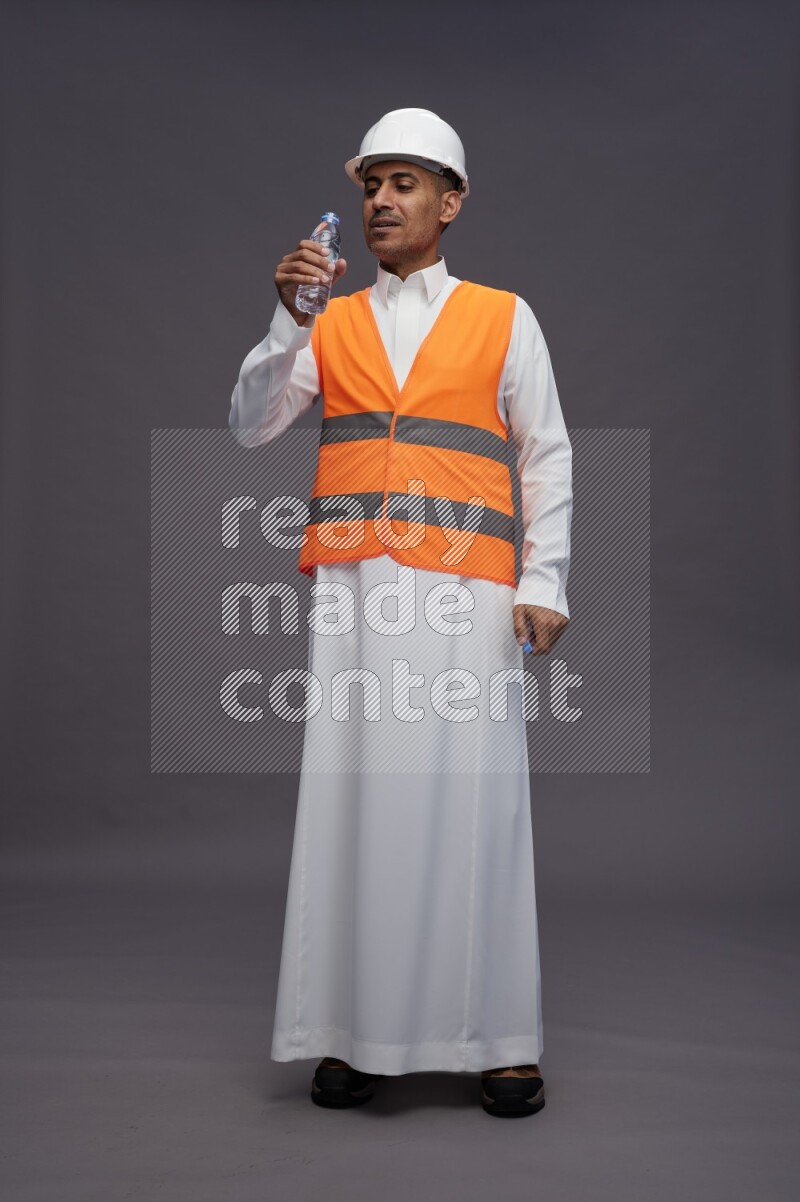 Saudi man wearing thob with engineer vest standing drinking water on gray background