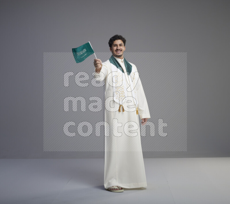 A Saudi man standing wearing thob and saudi flag scarf and holding small saudi flag on gray background