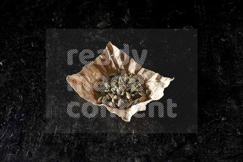 Cardamom seeds in a crumpled piece of paper on textured black flooring