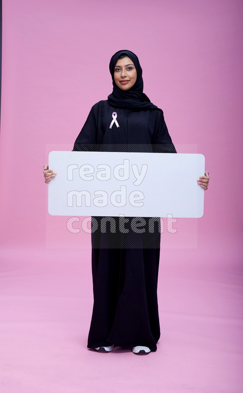 Saudi woman wearing pink ribbon on Abaya standing holding board on pink background