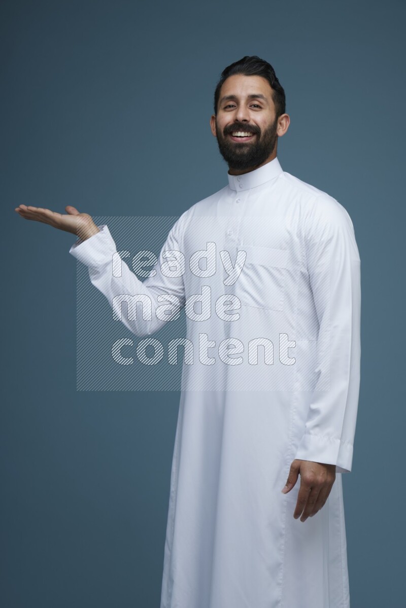 A man pointing in a blue background wearing Saudi Thob