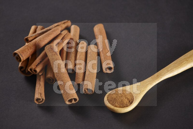 Cinnamon sticks stacked beside a wooden spoon full of cinnamon powder on black background