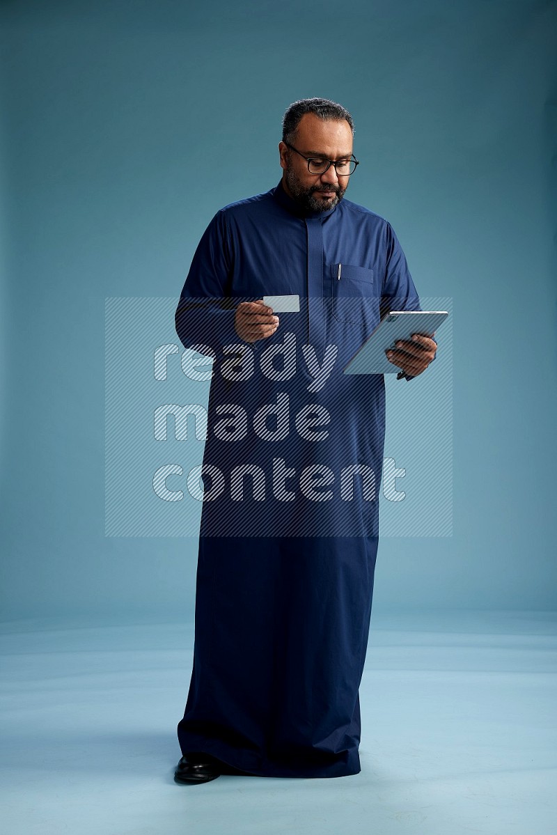 Saudi Man without shimag Standing holding ATM card while working on tablet on blue background
