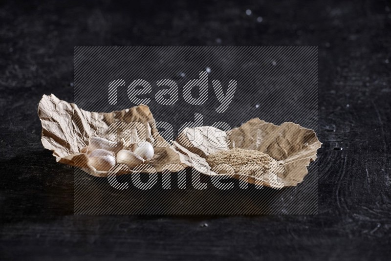2 crumpled piece of paper full of garlic cloves and powder on a textured black flooring in different angles