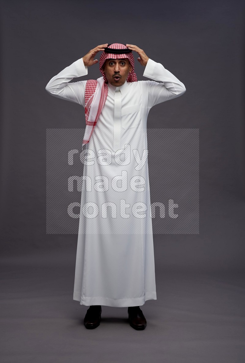 Saudi man wearing thob and shomag standing hands behind head on gray background