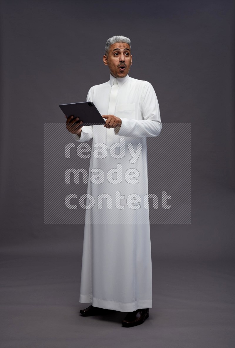 Saudi man wearing thob standing working on tablet on gray background