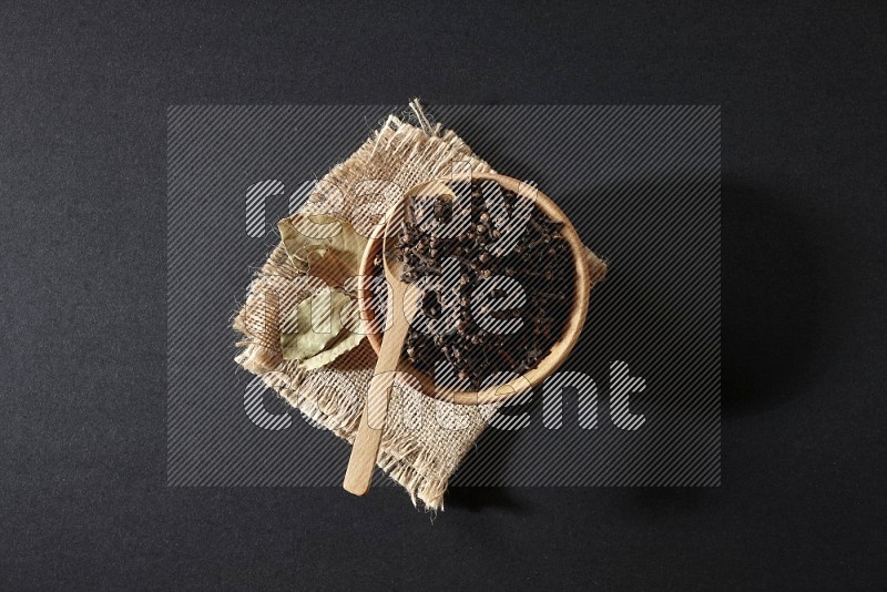 A wooden bowl, a wooden spoon full of cloves, and bay leaves (laurel) on a piece of burlap on a black flooring