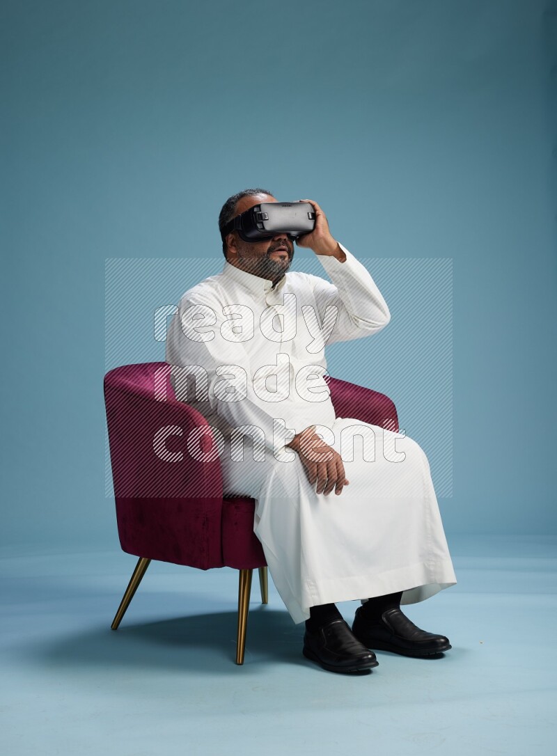 Saudi Man without shimag sitting on chair  gaming with VR on blue background