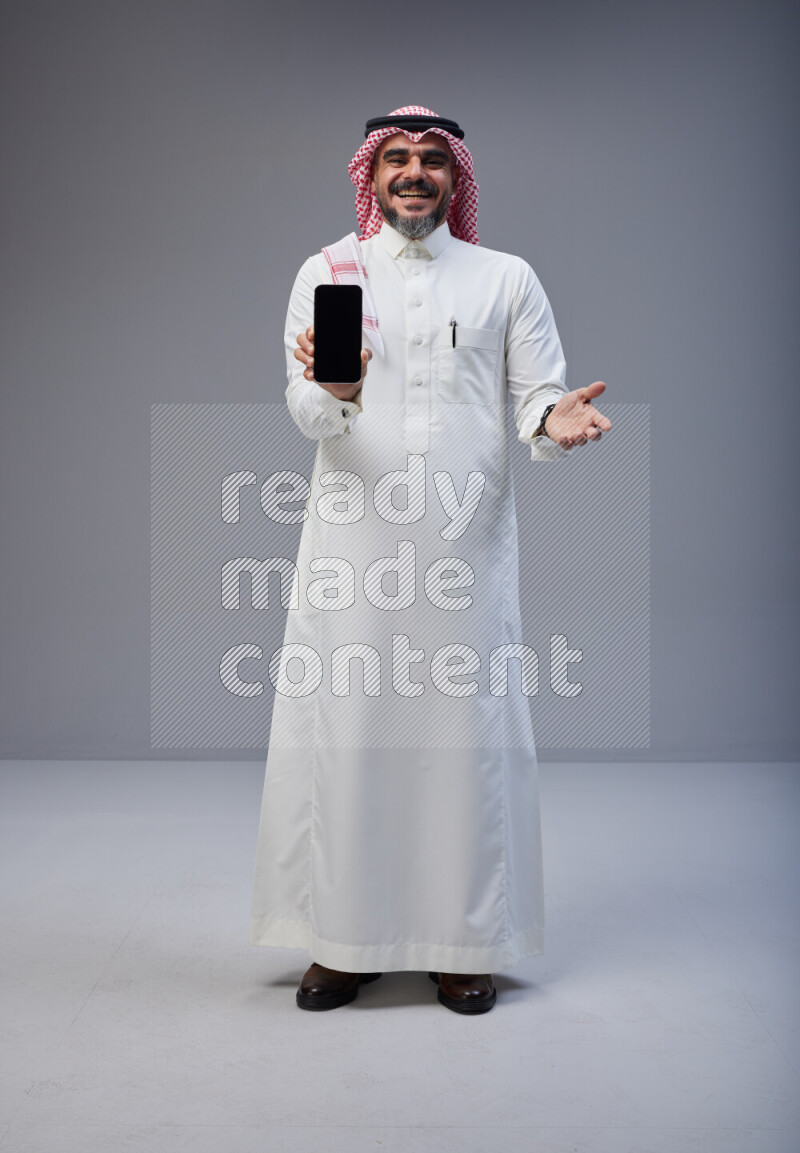 Saudi man Wearing Thob and red Shomag standing showing phone to camera on Gray background