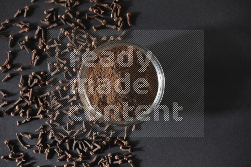 وعاء زجاجي ممتلئ ببودرة القرنفل مع حبوب القرنفل المتناثرة على أرضية سوداء