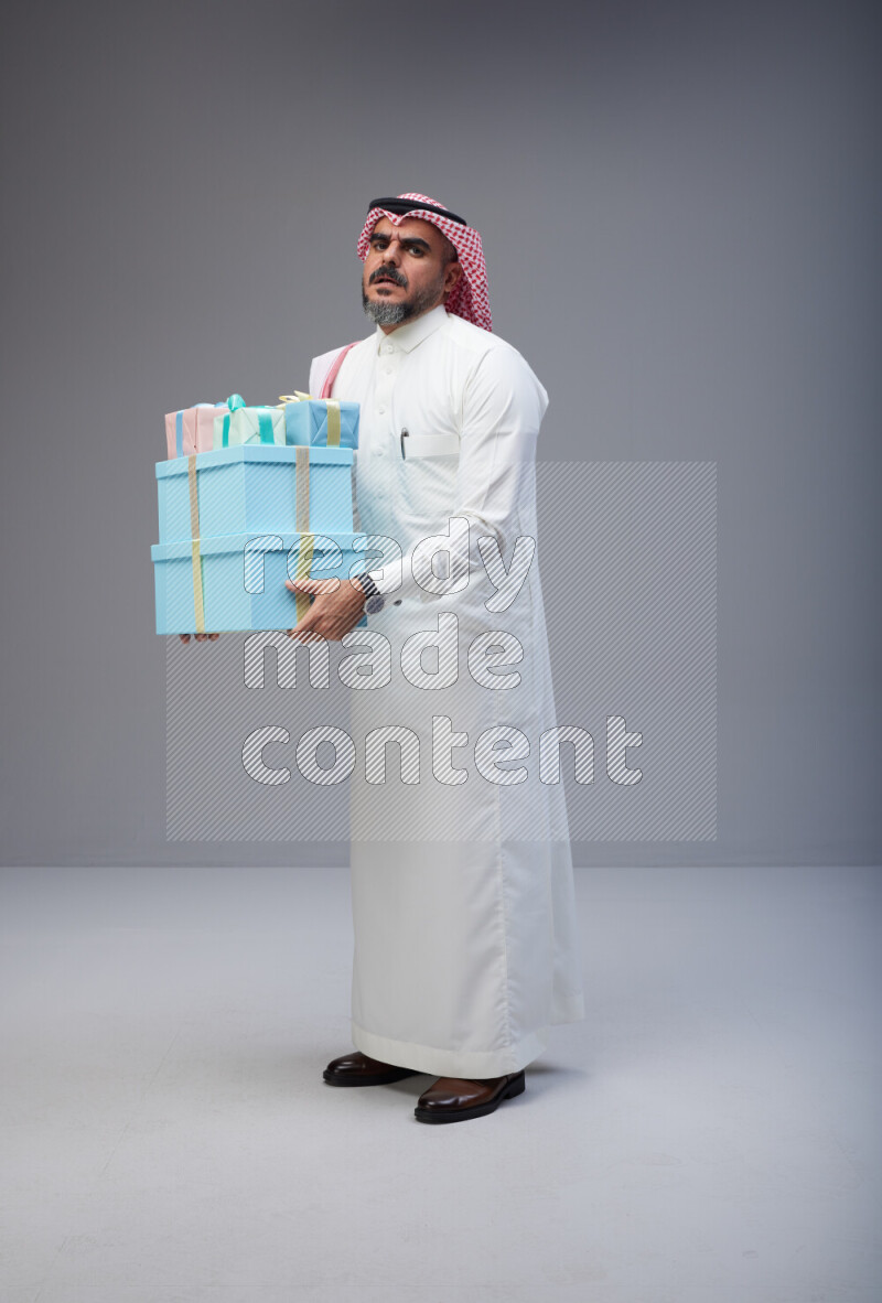 Saudi man Wearing Thob and red Shomag standing holding gift box on Gray background