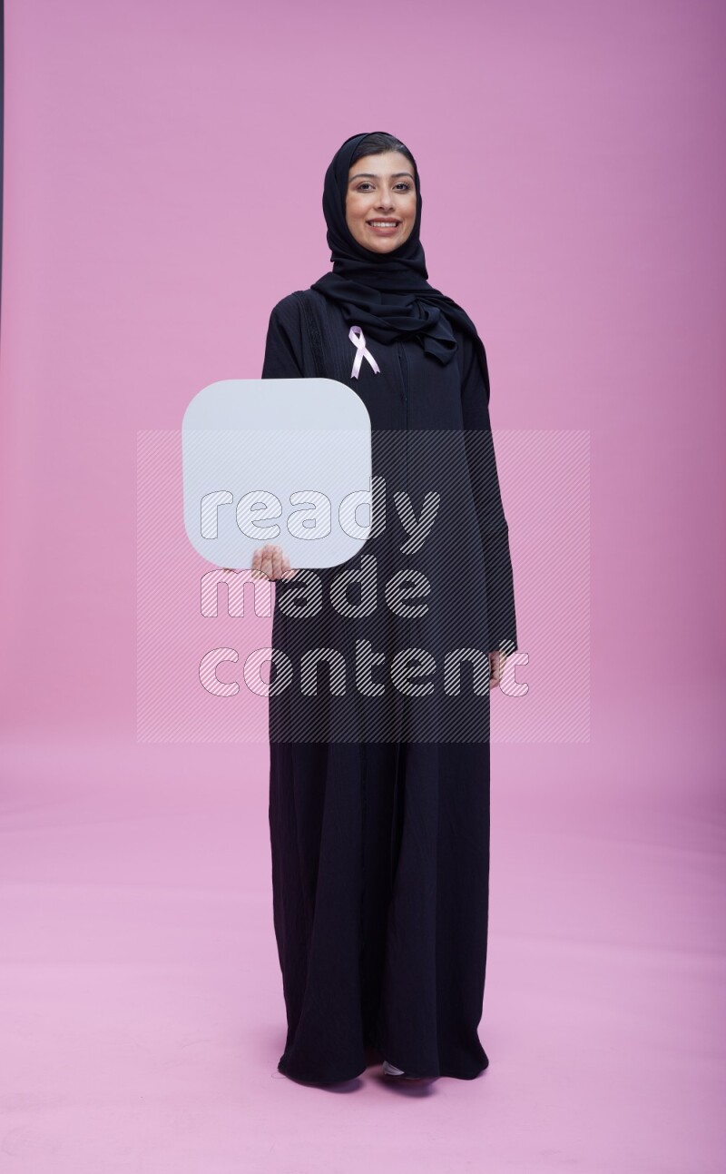 Saudi woman wearing pink ribbon on Abaya standing holding social media signs on pink background