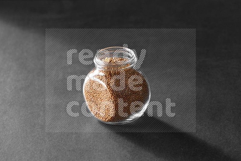 A glass spice jar full of mustard seeds on black flooring