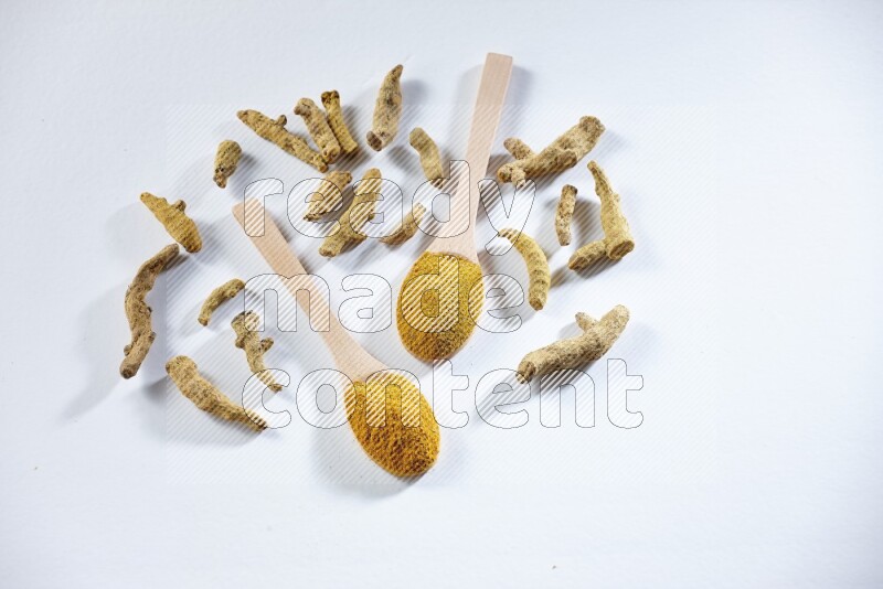 2 wooden spoons full of turmeric powder with dried turmeric fingers beside it on white flooring