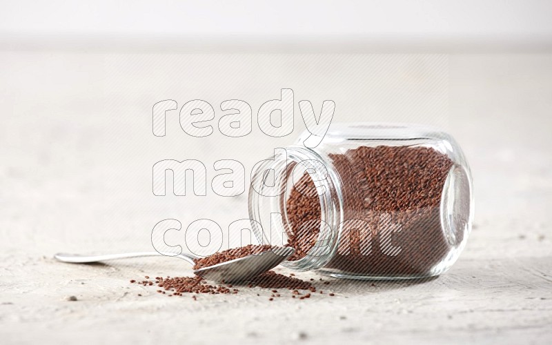 A glass spice jar and a metal spoon full of garden cress seeds and jar is flipped with fallen seeds on a textured white flooring