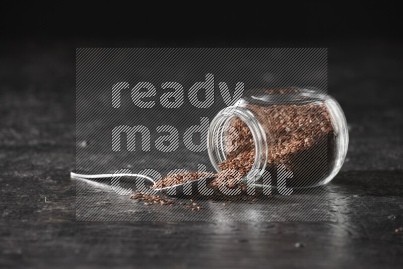 A glass spice jar full of flaxseeds flipped and seeds spread out with a metal spoon full of the seeds on a textured black flooring