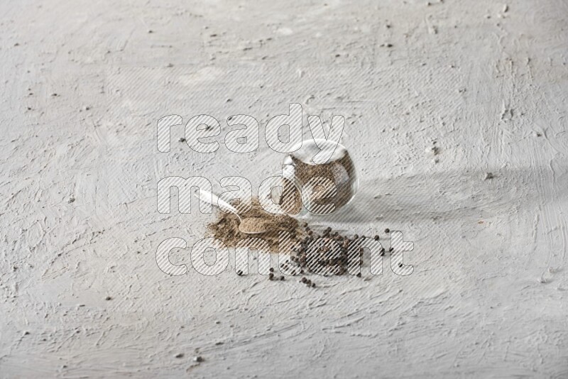 Flipped glass spice jar full of black pepper powder with a metal spoon and black pepper beads spread on a textured white flooring