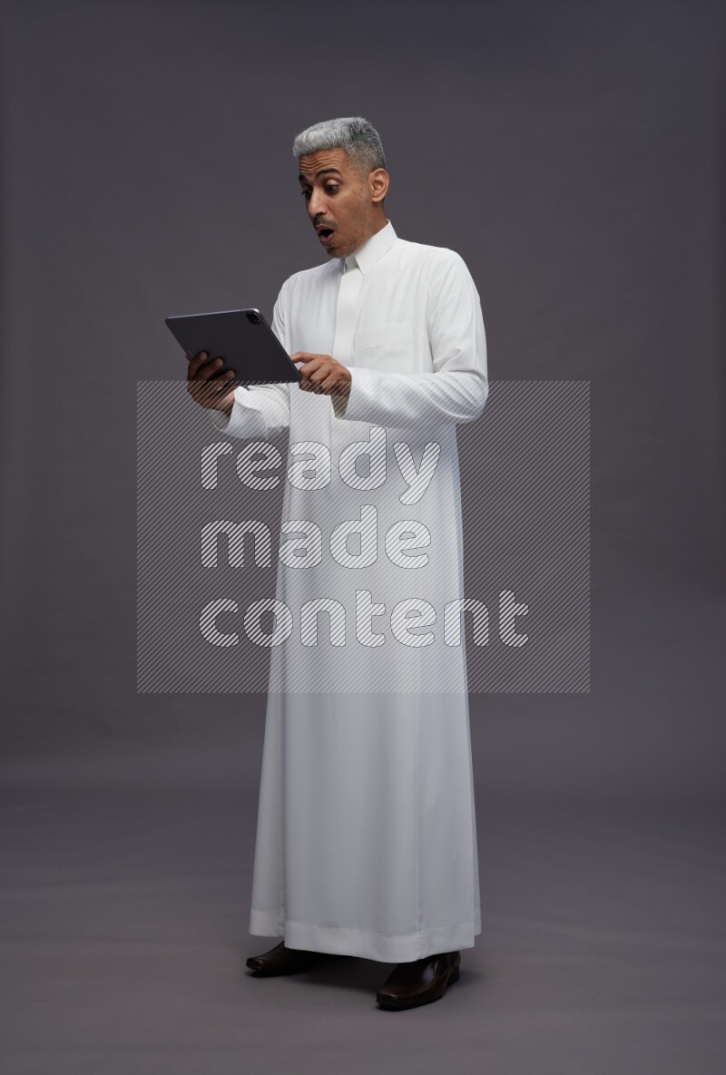 Saudi man wearing thob standing working on tablet on gray background