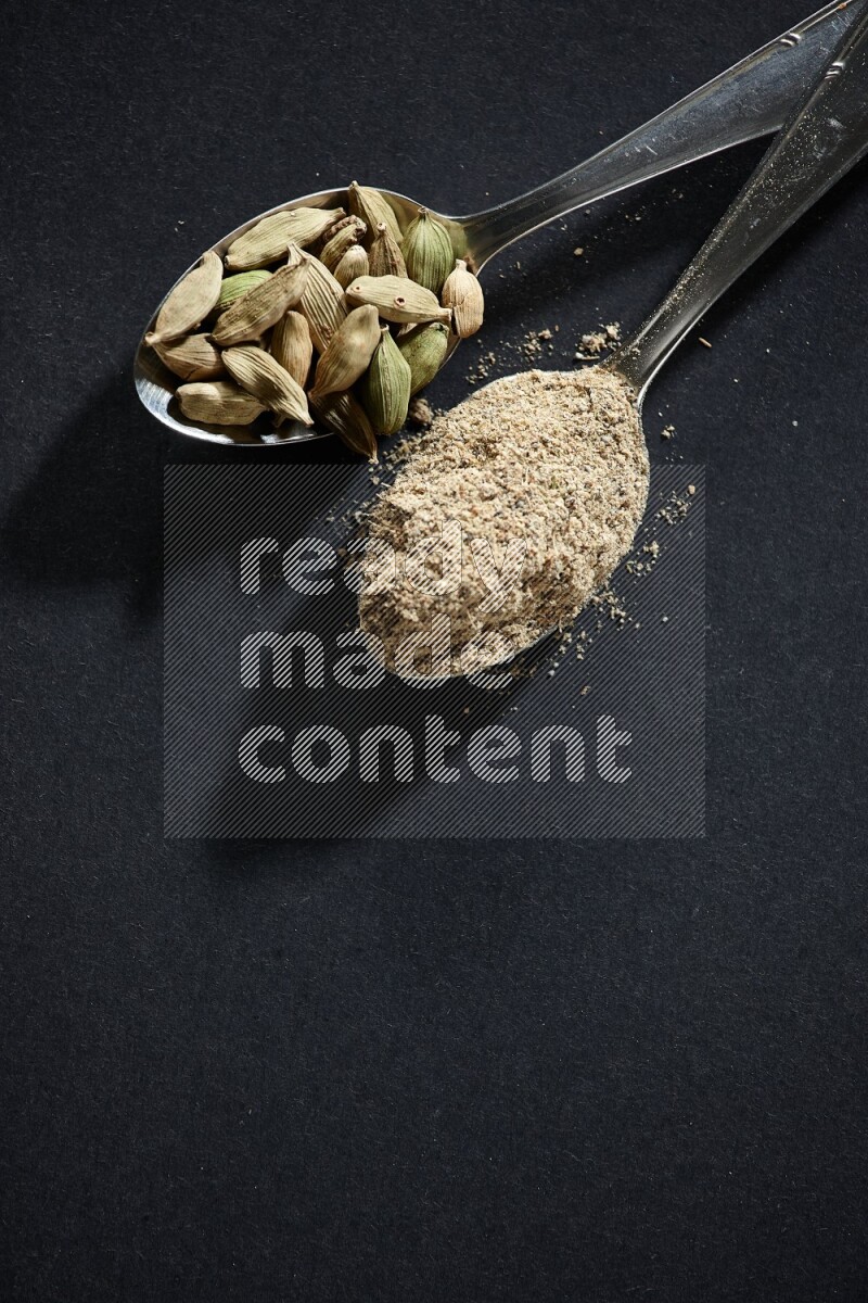 2 Metal spoons full of cardamom powder and cardamom seeds on black flooring