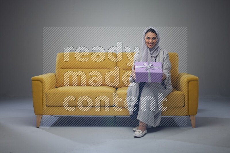 A Saudi woman wearing a light gray Abaya and head scarf sitting on a yellow sofa holding a gift box beside multi color gift boxes eye level on a grey background