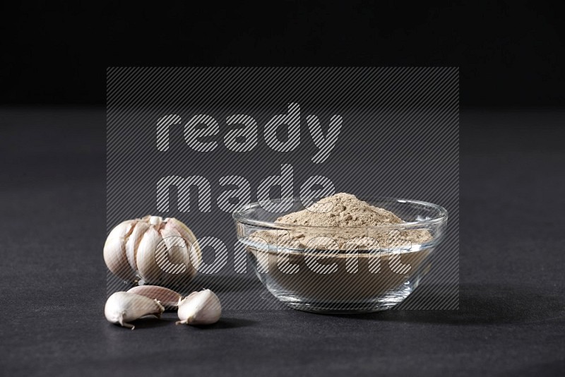A glass bowl full of garlic powder with garlic bulb and cloves beside it on a black flooring in different angles