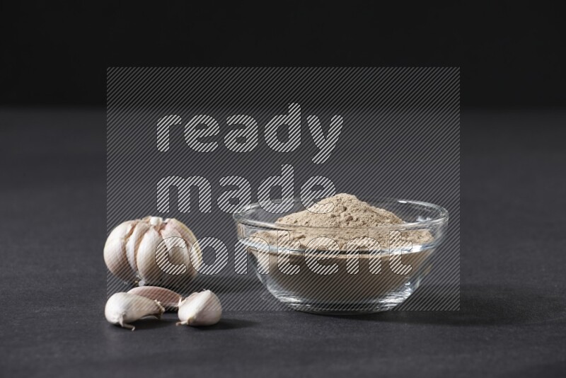 وعاء زجاجي ممتلئ ببودرة الثوم مع بعض فصوص الثوم ورأس ثوم كاملة على خلفية سوداء